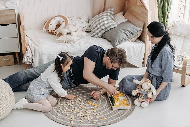 In einem Kinderzimmer sitzen ein Vater und eine Mutter mit ihrer Tochter auf dem Teppichboden und spielen mit Buchstabensteinen.