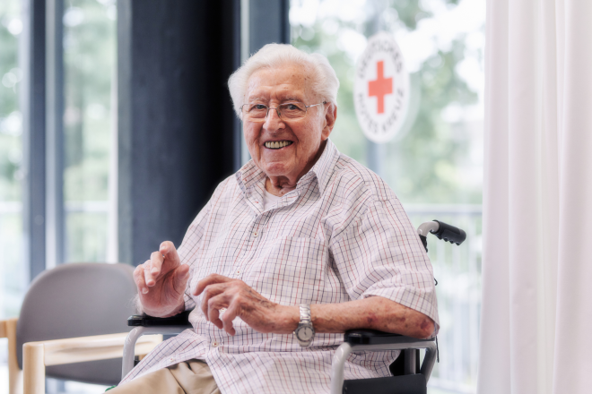 Bewohner sitzt auf einem Rollator und lächelt in die Kamera. Im Hintergrund ist an der Fensterscheibe das DRK-Logo zu sehen. 